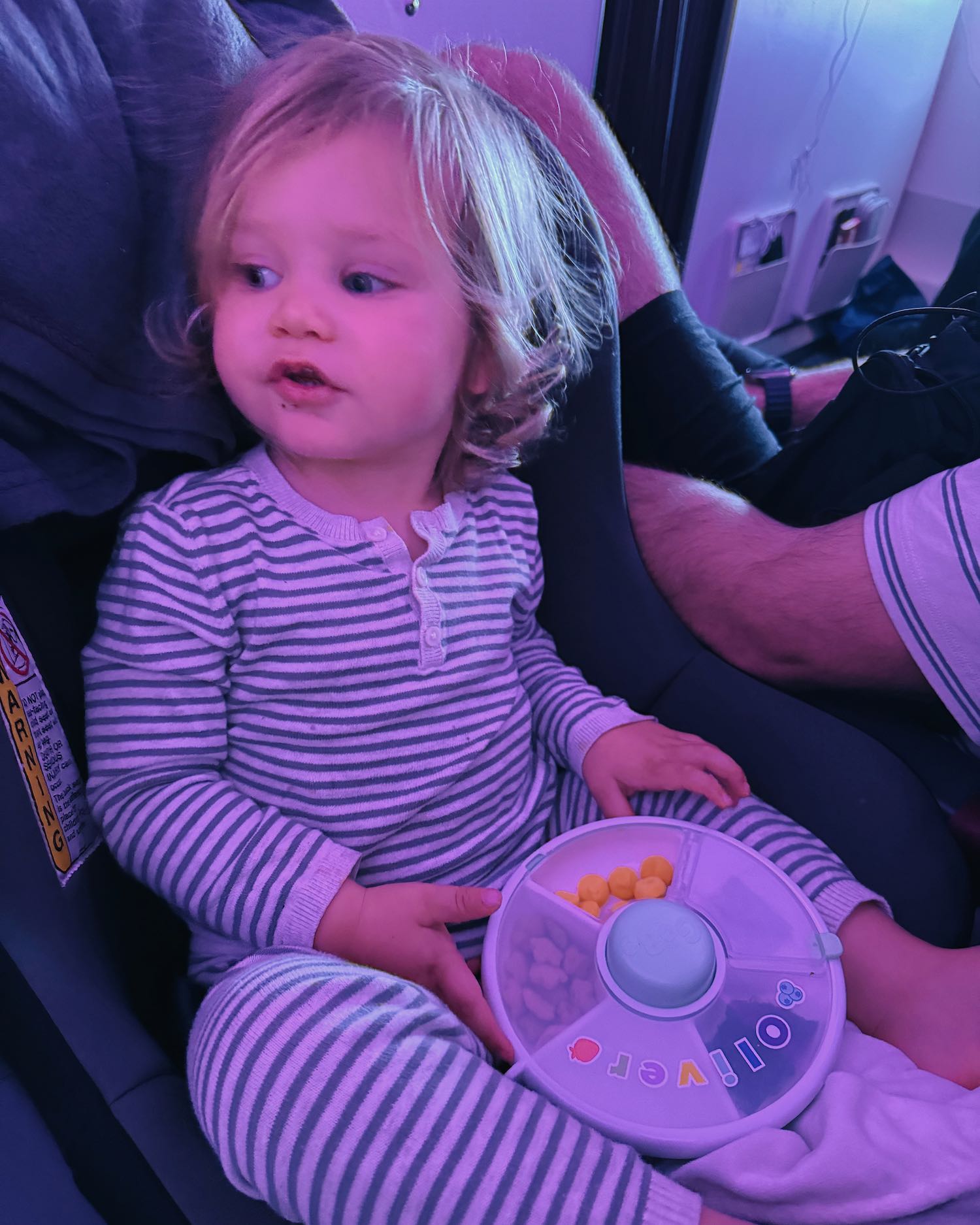 Blonde toddler in striped pajamas eating snacks out of snack box on airplane