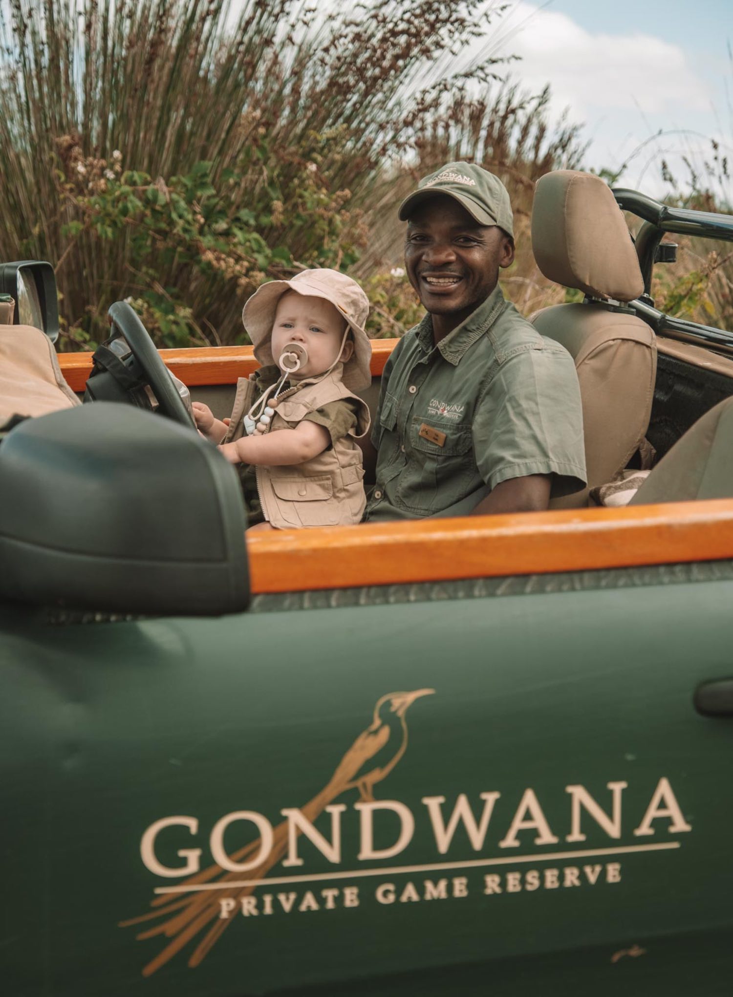 Man in olive green uniform smiling at camera sitting in driver's seat of jeep with baby in matching olive and khaki with pacifier in mouth and hands on steering wheel in Jeep that says GONDWANA PRIVATE GAME RESERVE on door