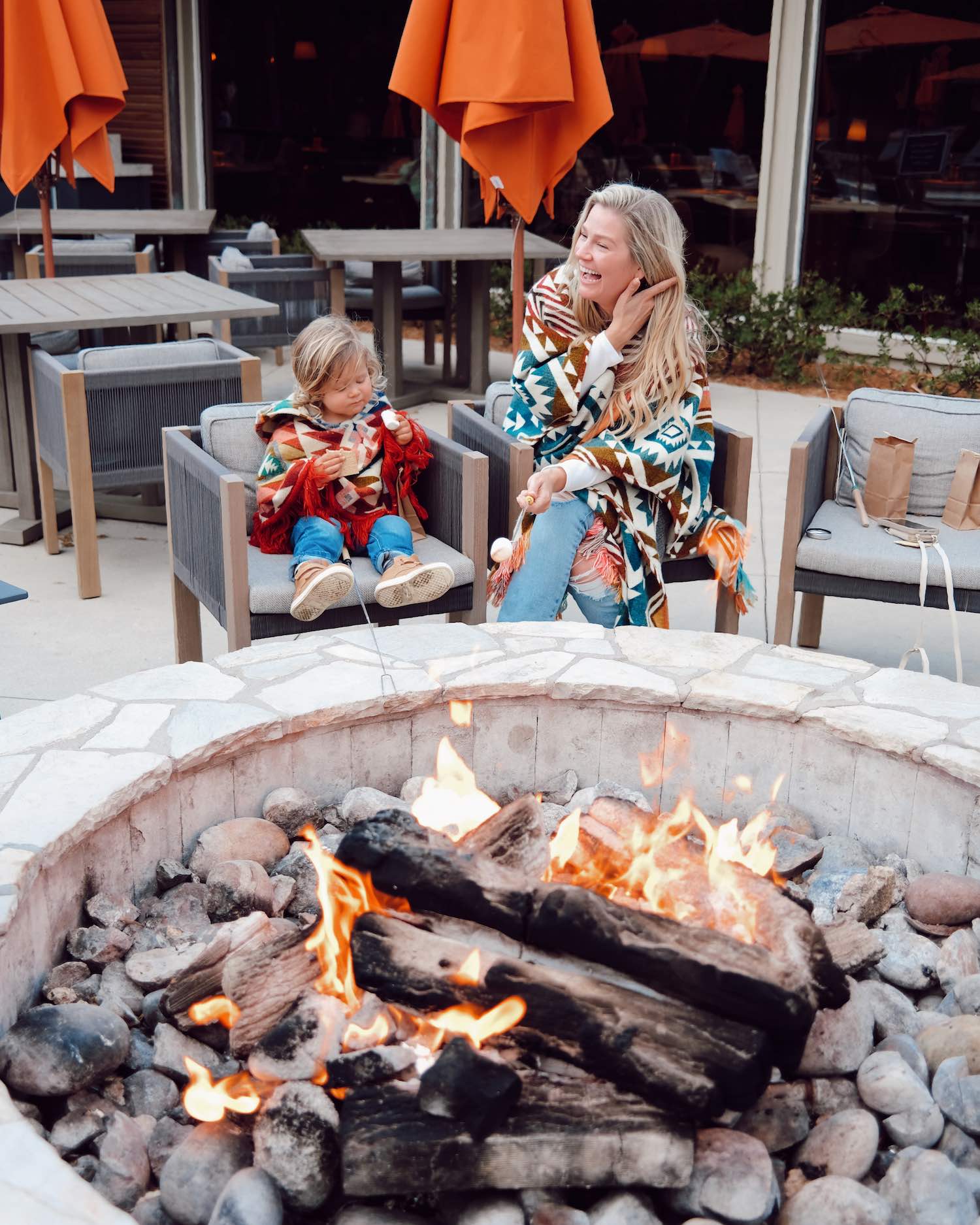 Woman laughing as she holds a marshmallow over an open flame with little toddler boy eating marshmallow beside her with fire in foreground