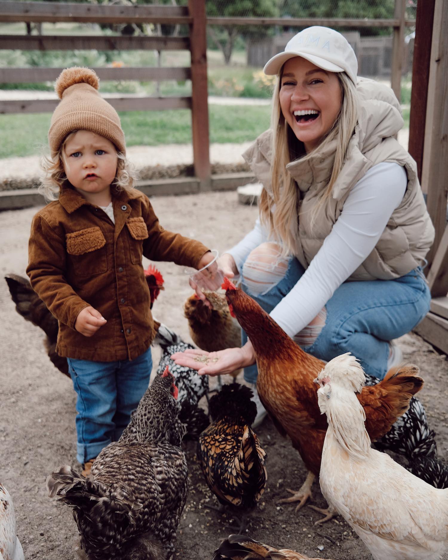 Woman in white shirt and beige puffer vest smiling off camera crouching down with toddler boy in brown long sleeve shirt and beanie feeding chickens at farm