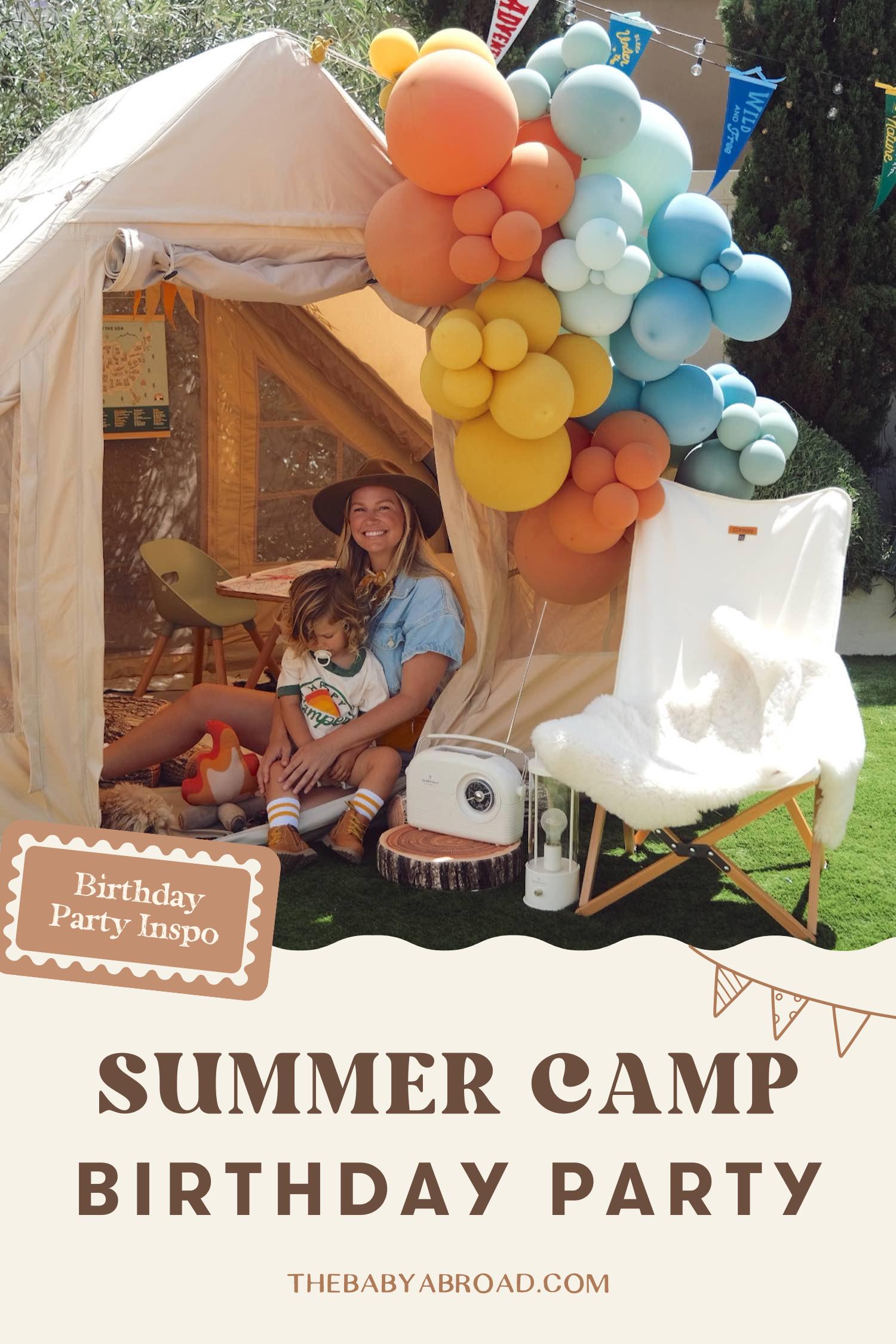 Woman with blue blouse and wide brimmed brown hat sitting in tent holding toddler boy with multi sized colorful balloons to the right, a lounge chair with fur and retro radio on a log slice on the ground