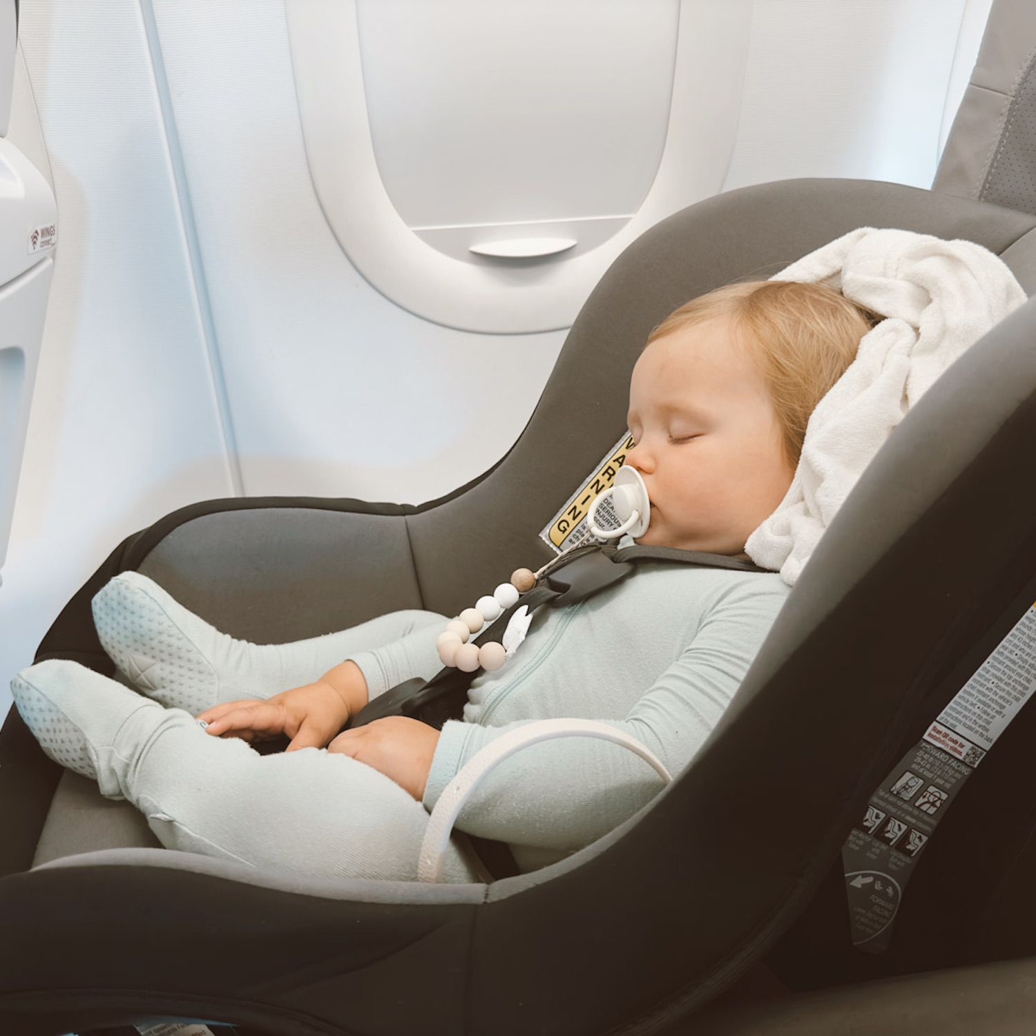 Small baby with pacifier in mouth sleeping in baby carrier in blue onesie in window seat of plane