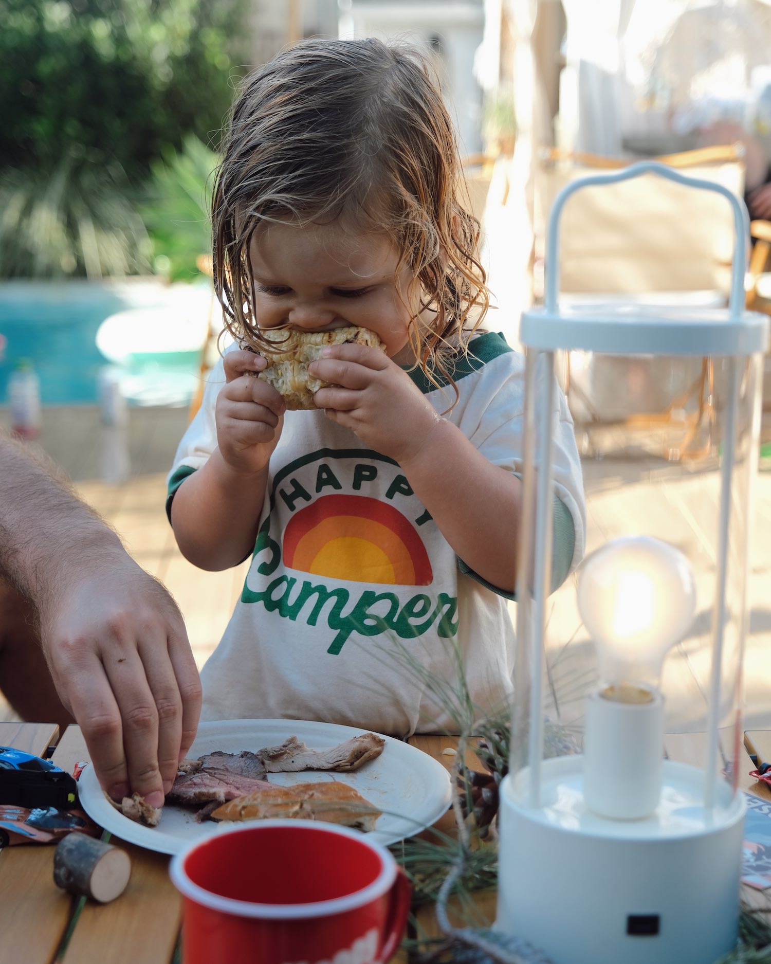 Little toddler boy with wet hair wearing a white tee with green Happy Camper letters on it using two hands to eat sandwich