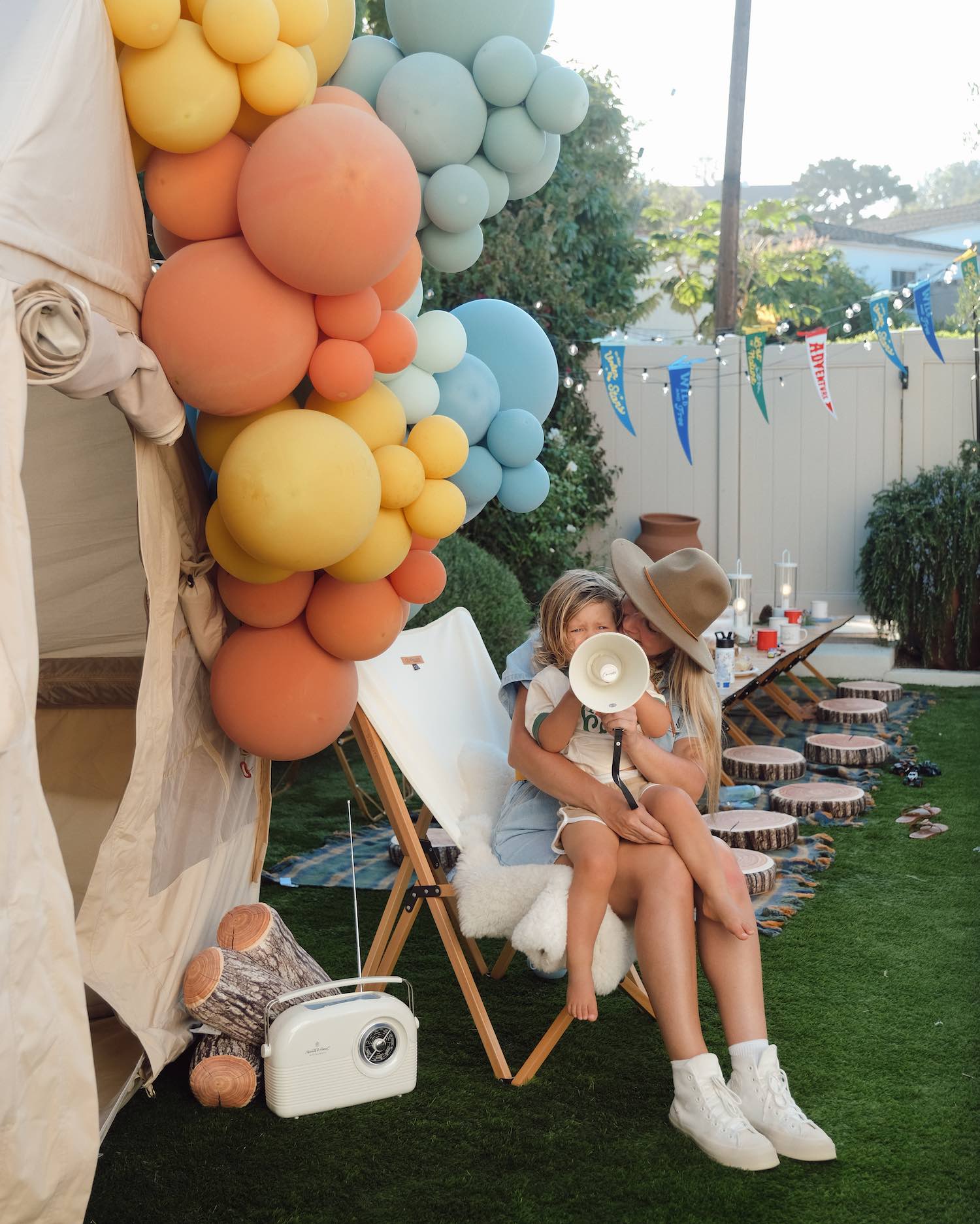 Woman with blue romper and wide brimmed brown hat sitting sitting in chair outside of tent holding little toddler boy holding up a megaphone to baby's face with multi sized colorful balloons above them, and a retro radio with faux logs decorated around it.