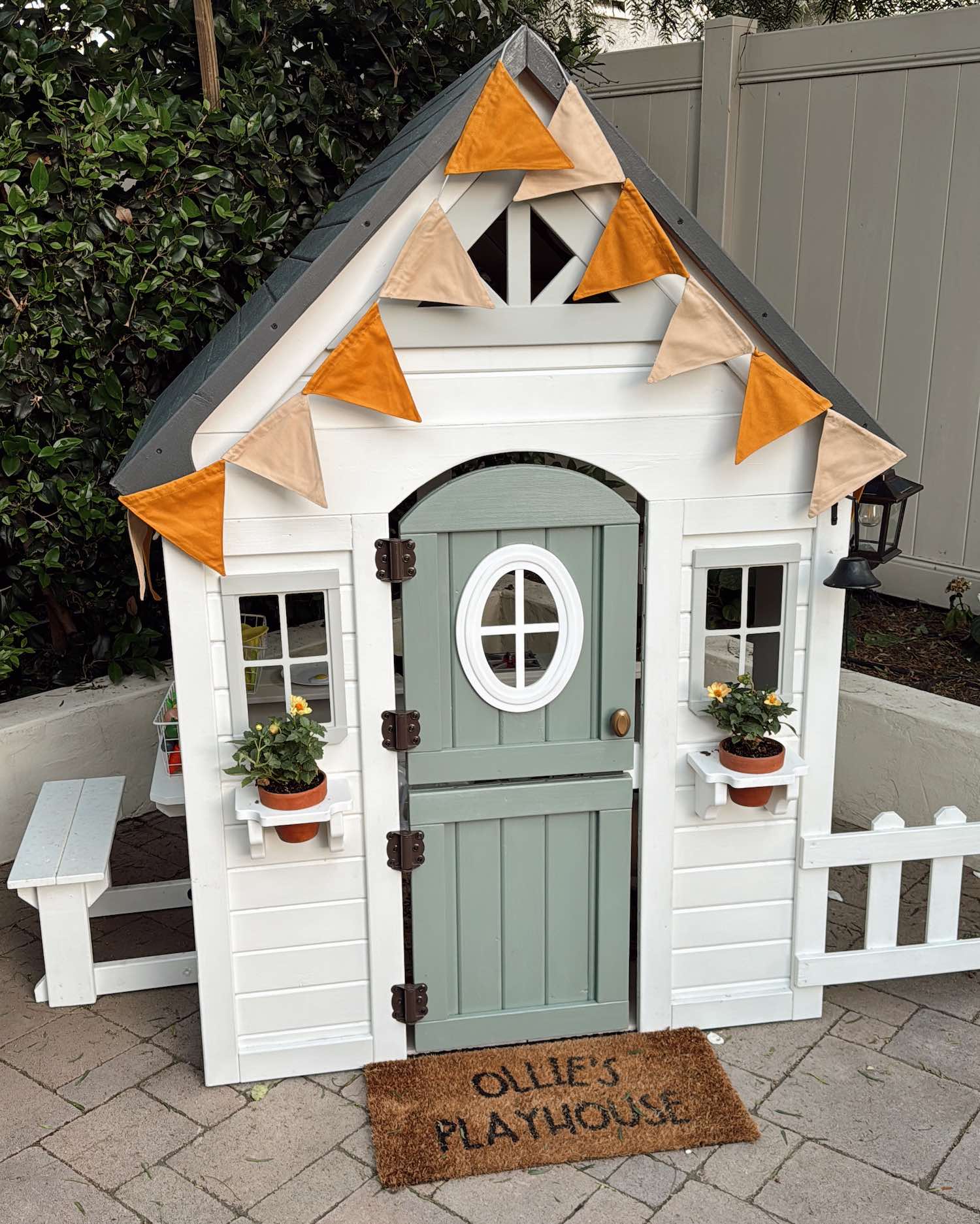 Small white child's playhouse with sage green door with yellow and orange flag banner along the top roof