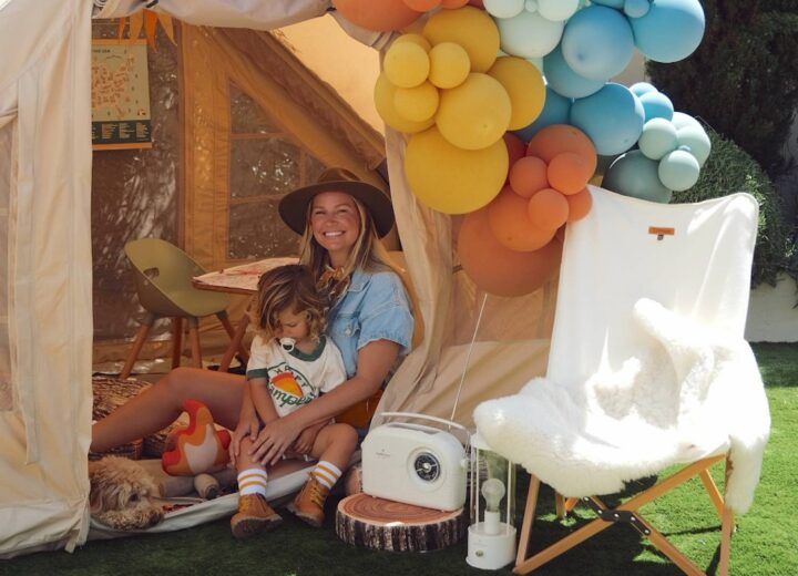 Woman with blue romper and wide brimmed brown hat sitting in tent holding toddler boy with multi sized colorful balloons to the right, a lounge chair with fur and retro radio on a log slice on the ground