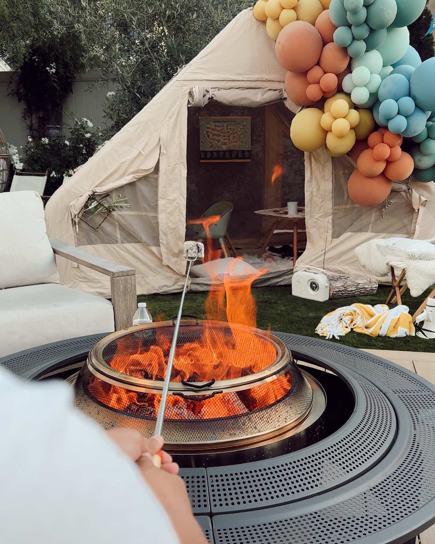 Person holding marshmallow on a skewer over open flame on outdoor stove with tent and balloons in the background