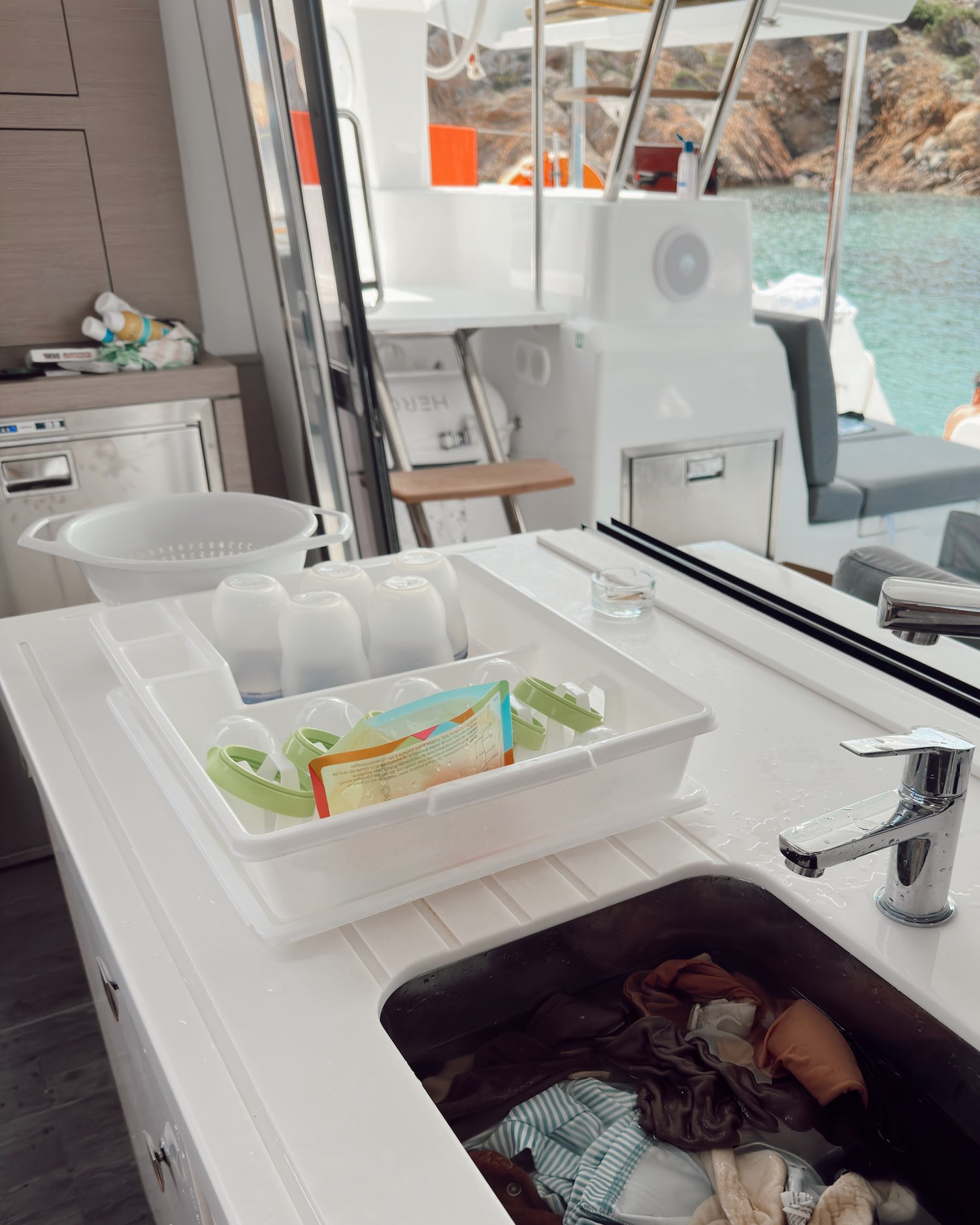 Kitchen sink full of clothes and bathing suits in water with drying baby bottles and feeding supplies drying in plastic bin to the left