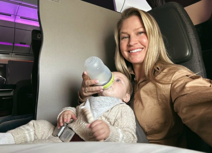 Blonde woman taking selfie in one hand with baby in another hand as she feeds him a bottle sitting in first class flight on Qatar Airways