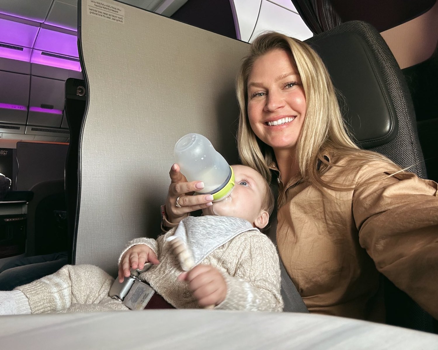 Blonde woman taking selfie in one hand with baby in another hand as she feeds him a bottle sitting in first class flight on Qatar Airways