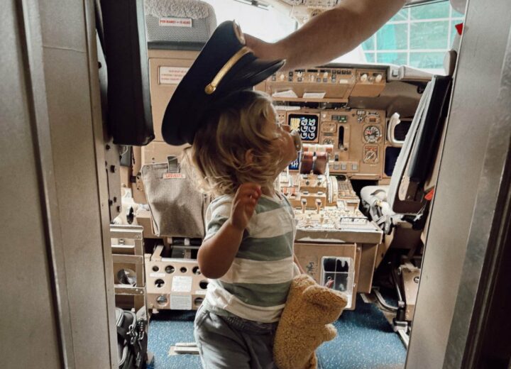 blonde toddler with pacifier in stripes with his teddy bear looking at out-of-frame pilot as pilot places captain's hat on toddler in cockpit