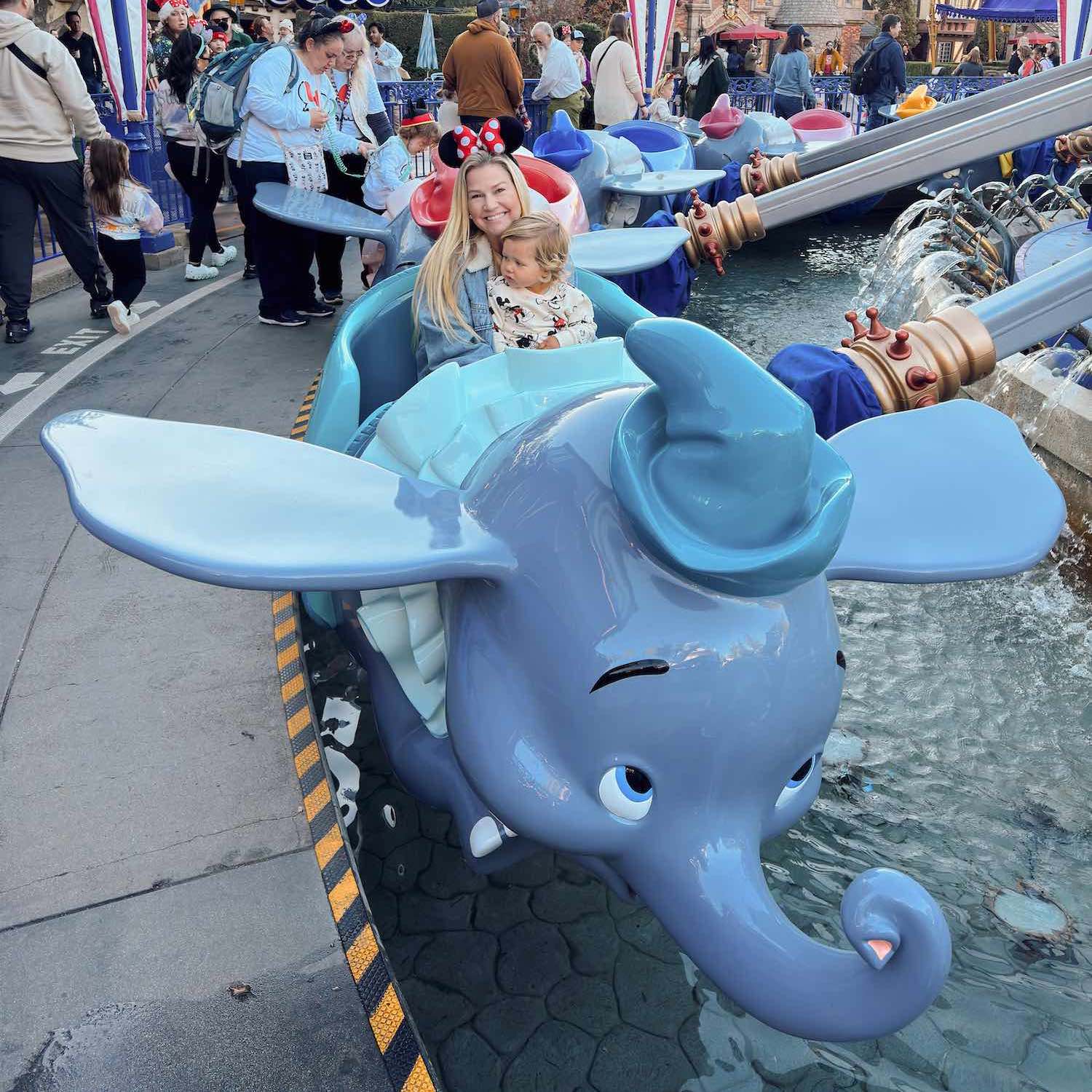 Woman with long blonde hair and Minnie Mouse Ears smiling at camera with little boy sitting in front of her as they sit in a Dumbo figure for the Flying Dumbo Ride at Fantasyland in Disney