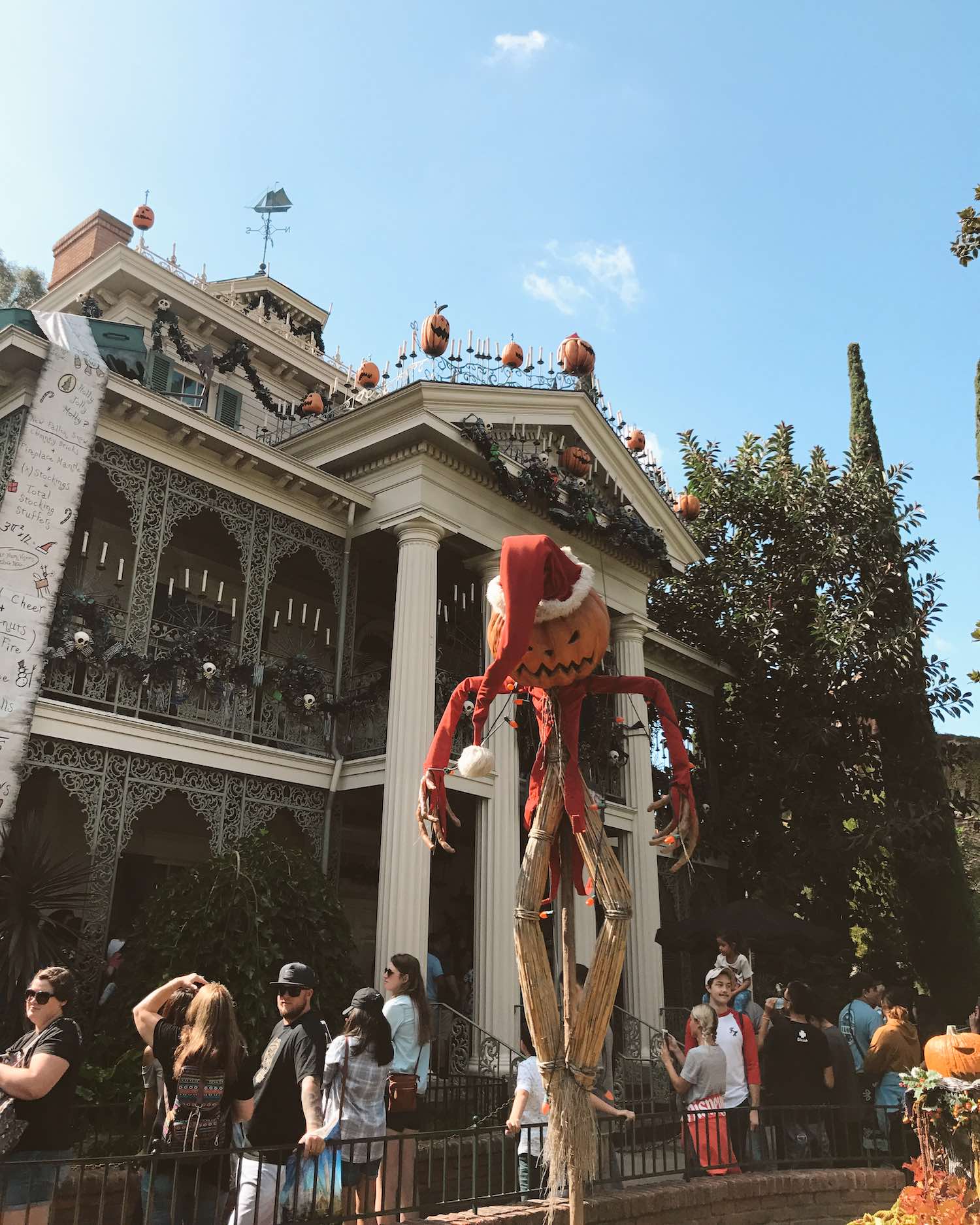 Scarecrow with Pumpkin head and Santa Clause outfit in front of the Haunted Mansion with a line of people waiting in queue