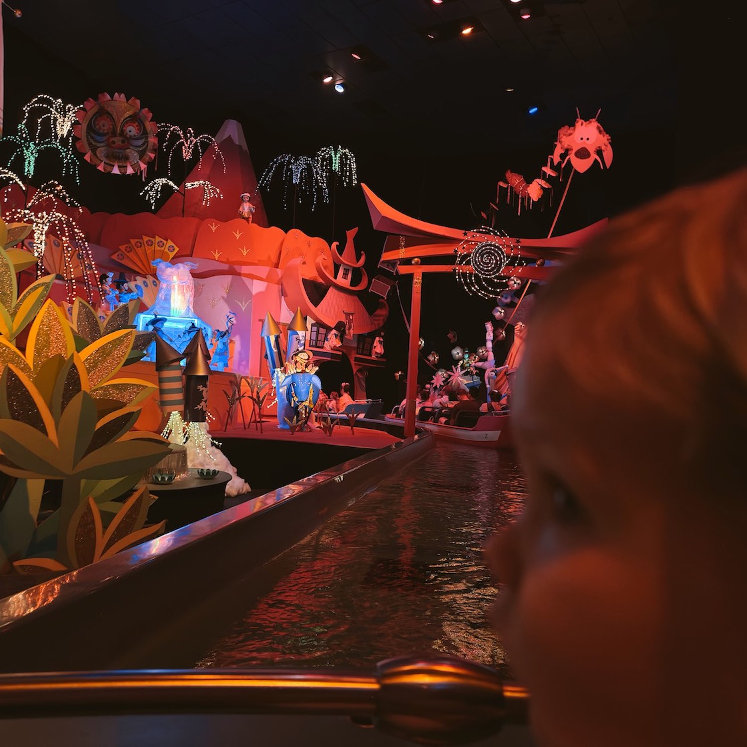 Little boy in front of camera blurry as he looks out of the boat from It's a Small World with the lit decor and amusement on the rides lit up